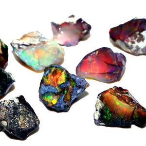 Puede incluir: Una colección de ocho piedras preciosas de ópalo en bruto de varios colores, incluyendo negro, verde, rojo y azul. Los ópalos son iridiscentes y tienen una variedad de formas y tamaños.