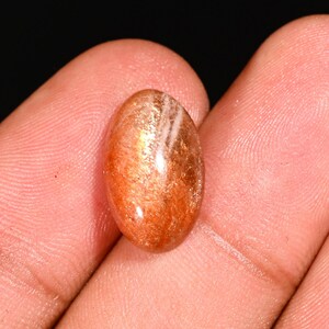 Könnte beinhalten: Ovaler Sonnenstein-Edelstein mit polierter Oberfläche. Der Stein hat eine warme orangebraune Farbe mit schimmernden Einschlüssen und weißen Streifen. Der Edelstein wird in einer Hand gehalten, um seine Größe und Form zu zeigen.