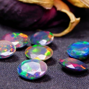 Könnte beinhalten: Sieben schillernde Opal-Edelsteine, jeder mit einem einzigartigen Farbmuster, werden auf einer dunklen Stoffoberfläche präsentiert. Die Opale sind oval geformt und haben einen facettierten Schliff.