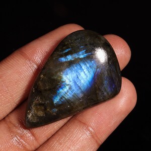 Puede incluir: Un cabujón de labradorita gris oscuro con destellos iridiscentes azules y verdes. La piedra es lisa y pulida, con forma de lágrima.