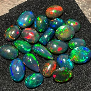 May include: A collection of black opal gemstones with iridescent green, blue, and red flashes. The opals are various shapes and sizes, including oval, pear, and round.