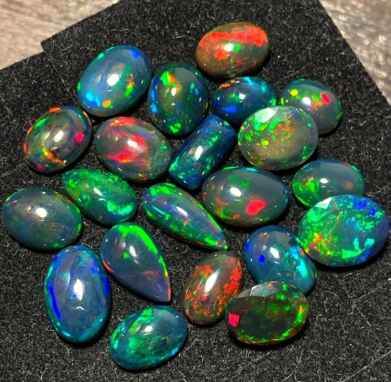 May include: A collection of black opal gemstones with iridescent green, blue, and red flashes. The opals are various shapes and sizes, including oval, pear, and round.