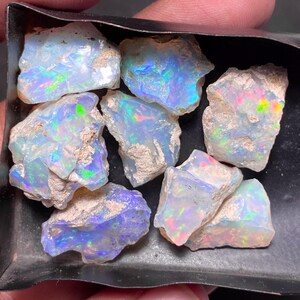 May include: A collection of rough opal gemstones, showcasing a play of color with iridescent flashes of blue, green, and red. The opals have a raw, natural appearance, with a mix of milky white and earthy brown tones. The gemstones are displayed in a dark tray.