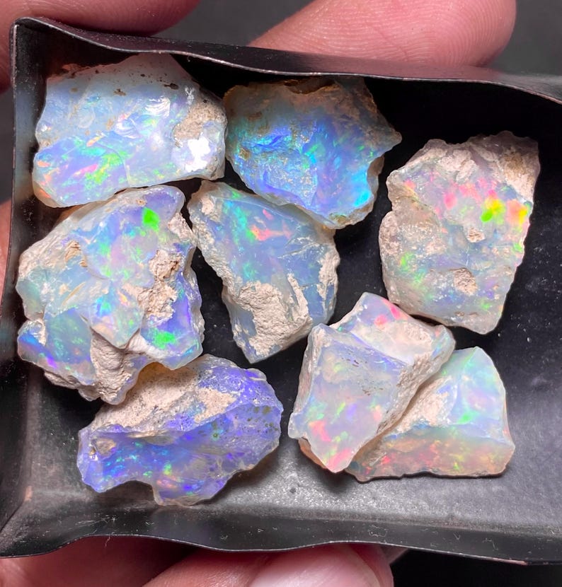 May include: A collection of rough opal gemstones, showcasing a play of color with iridescent flashes of blue, green, and red. The opals have a raw, natural appearance, with a mix of milky white and earthy brown tones. The gemstones are displayed in a dark tray.
