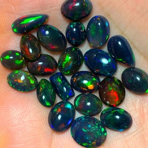 May include: A collection of black opal gemstones in various oval and teardrop shapes. The opals display a range of iridescent colors, including green, blue, red, and gold.