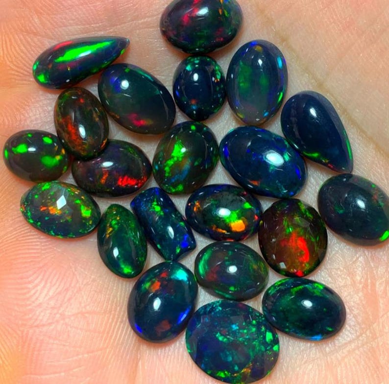 May include: A collection of black opal gemstones in various oval and teardrop shapes. The opals display a range of iridescent colors, including green, blue, red, and gold.