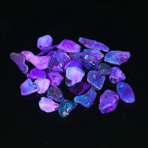 May include: A collection of raw opal gemstones in shades of purple, blue, and pink. The opals have a polished, iridescent surface, with flashes of color. The gemstones are scattered on a black surface, creating a striking contrast.