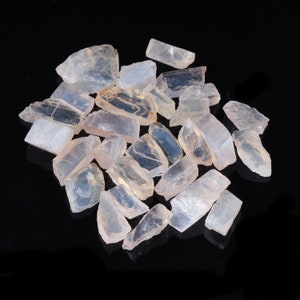 May include: A collection of translucent, pale pink and white selenite crystal chunks. The crystals have irregular shapes and varying sizes, scattered on a reflective black surface. The light catches the facets, creating highlights and shadows.