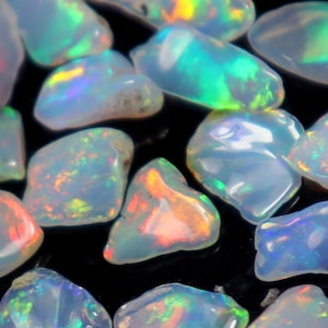 Könnte beinhalten: Eine Sammlung von regenbogenfarbenen Opal-Edelsteinen in verschiedenen Formen und Größen. Die Opale sind milchig weiß mit grün, blau und roten Schimmern.