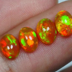 Könnte beinhalten: Vier ovale Edelsteine mit einem leuchtenden orangefarbenen, grünen und gelben irisierenden Schimmer. Die Edelsteine sind in einer Reihe auf einem Finger angeordnet.