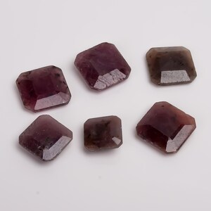 Puede incluir: Seis piedras preciosas facetadas en tonos morados y marrones están dispuestas sobre una superficie blanca. Las piedras tienen forma cuadrada o rectangular con bordes biselados. Las piedras varían en color, desde el morado intenso hasta un tono parduzco.