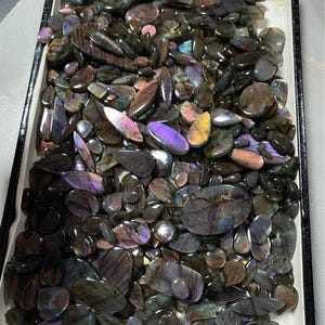 May include: A tray filled with polished labradorite gemstones. The stones vary in shape and size, displaying a range of colors including gray, brown, purple, and blue. The gemstones are arranged in a rectangular tray with a black border.