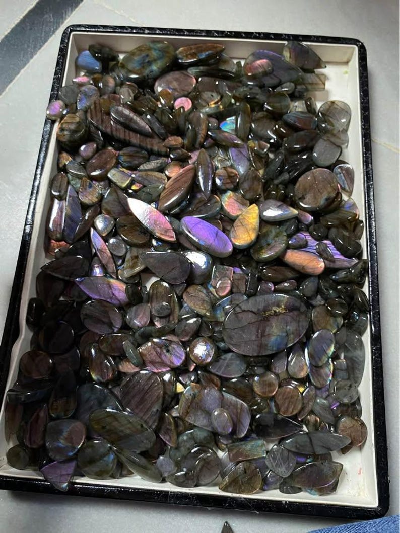 May include: A tray filled with polished labradorite gemstones. The stones vary in shape and size, displaying a range of colors including gray, brown, purple, and blue. The gemstones are arranged in a rectangular tray with a black border.