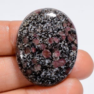 Może przedstawiać: Owalny kamień szlachetny w szlifie kaboszonowym z czarno-białym wzorem w kropki i małymi, rozproszonymi różowymi wtrąceniami.