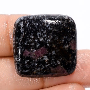 Peut inclure: Une pierre précieuse cabochon noire et blanche avec des inclusions roses et violettes. La pierre précieuse est de forme carrée et a une surface lisse et polie.