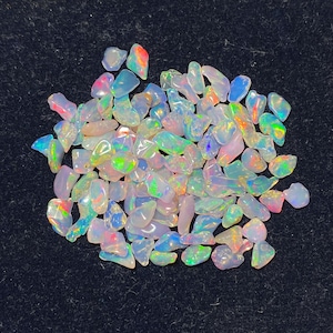 May include: A collection of small, tumbled opal gemstones featuring a translucent, milky white base with vibrant flashes of iridescent color, including neon green, bright orange, and electric blue, scattered across a dark, textured surface.