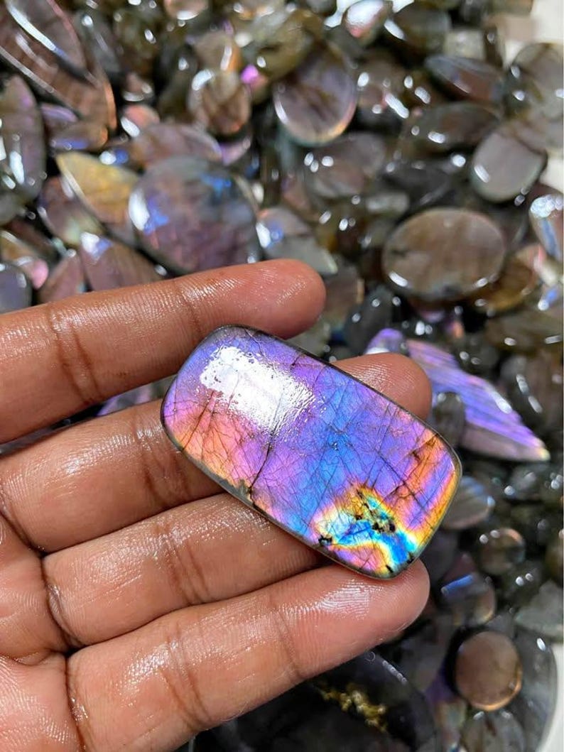 May include: A rectangular, polished labradorite gemstone with a vibrant display of colors. The stone features a mix of purple, blue, and yellow hues, with a rainbow-like effect. The gemstone is held in a hand, with other similar stones in the background.