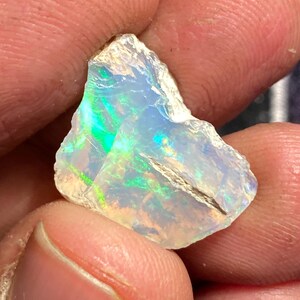 May include: A rough-cut opal gemstone, displaying a play of color with iridescent flashes of green, blue, and yellow. The stone is held between fingers, showcasing its natural, irregular shape and translucent quality.