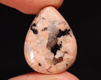 May include: A pink and white teardrop-shaped cabochon with black and green inclusions. The cabochon is smooth and polished.