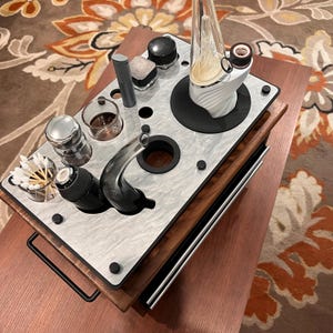 Puede incluir: Una bandeja organizadora rectangular de madera con una superficie superior de mármol plateado que presenta múltiples recortes circulares. La bandeja contiene un vaporizador de vidrio blanco y negro, una pipa de vidrio negro, un frasco de vidrio pequeño, una herramienta de metal y una taza de vidrio con hisopos de algodón.