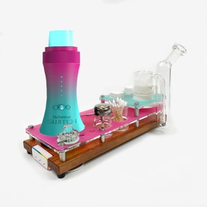 Könnte beinhalten: Ein rosa-blauer Dr. Dabber Switch Vaporizer mit einer Glaswasserpfeife, einem kleinen Glasgefäß, Wattestäbchen und einem Holzsockel. Der Vaporizer hat ein digitales Display und einen Knopf.