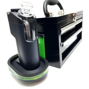 May include: A black electronic device with a clear glass attachment sits on a circular base with a green accent stripe. Next to it is a black metal tool chest with a handle and silver drawer pulls, featuring the word Husky printed on the front.