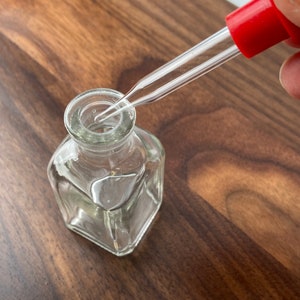 May include: A clear glass bottle with a square base and a narrow neck. A glass dropper with a red rubber top is positioned above the bottle's opening, ready to dispense liquid. The bottle sits on a dark wood surface.