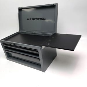 May include: A gray U.S. General metal tool chest with an open lid, featuring two drawers and a black side shelf. The interior of the lid displays the U.S. General logo in black text, and the front of the chest also features the U.S. General brand name.