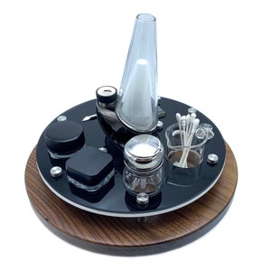 May include: A black and silver glass bong with a wooden base. The bong is surrounded by small containers and a glass jar with cotton buds.