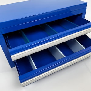 May include: A blue metal storage box with two open drawers. The drawers have silver trim and dividers. The interior of the box and drawers are blue. The box is designed for organizing small items.