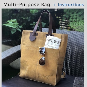 以下が含まれることがあります： 正面に大きなポケットが付いた茶色の革製のトートバッグ。籐製の椅子の上に置かれ、背景は緑の葉で覆われています。バッグのサイズは、幅31インチ、高さ40インチ、奥行き15インチです。