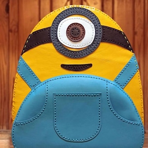 May include: A yellow leather Minion-themed backpack with a blue denim-style pocket on the front. The backpack has a single large eye with a black pupil and white iris. The eye is surrounded by black stitching.