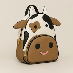 Può includere: Zaino a forma di mucca. Lo zaino presenta un corpo bianco con macchie nere, dettagli marroni e una faccia sorridente. Ha una maniglia superiore, spallacci regolabili e cerniere dorate.