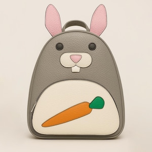 May include: Gray bunny-shaped backpack with pink-lined ears, black eyes, and a pink nose. The backpack features a white belly with an orange and green carrot design. A fun and functional accessory for kids.