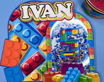 May include: A colorful cake topper with a blue, red, yellow, and green Lego-themed design. The topper features the name "IVAN" and the number "5" in a glittery, white background.