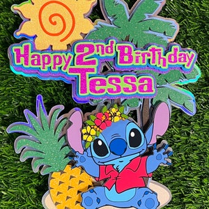 May include: A blue Stitch character cake topper with a red shirt and a pineapple. The topper is on a beach scene with a palm tree and a sun. The text "Happy 2nd Birthday Tessa" is on the topper.