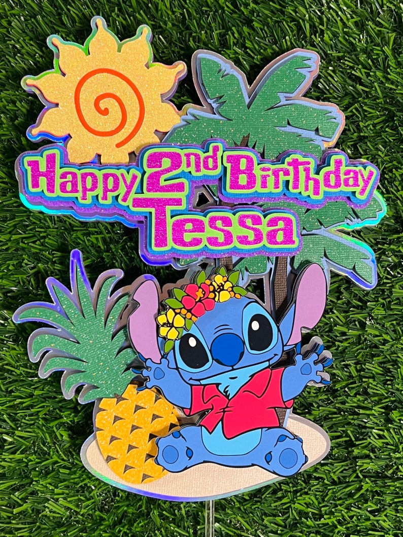 May include: A blue Stitch character cake topper with a red shirt and a pineapple. The topper is on a beach scene with a palm tree and a sun. The text "Happy 2nd Birthday Tessa" is on the topper.