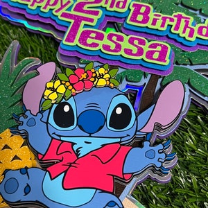 May include: A blue Stitch character wearing a red shirt and a flower crown. The character is standing on a beach scene with a pineapple and palm trees. The text "Happy 2nd Birthday Tessa" is in the background.