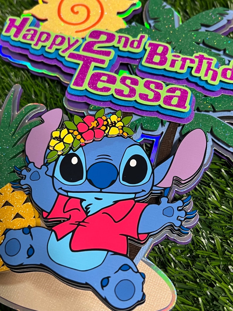May include: A blue Stitch character wearing a red shirt and a flower crown. The character is standing on a beach scene with a pineapple and palm trees. The text "Happy 2nd Birthday Tessa" is in the background.
