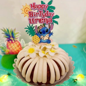 May include: A white cake with white frosting and a blue Stitch character cake topper. The cake topper also includes a pineapple, palm trees, and the text "Happy Birthday Lorena".