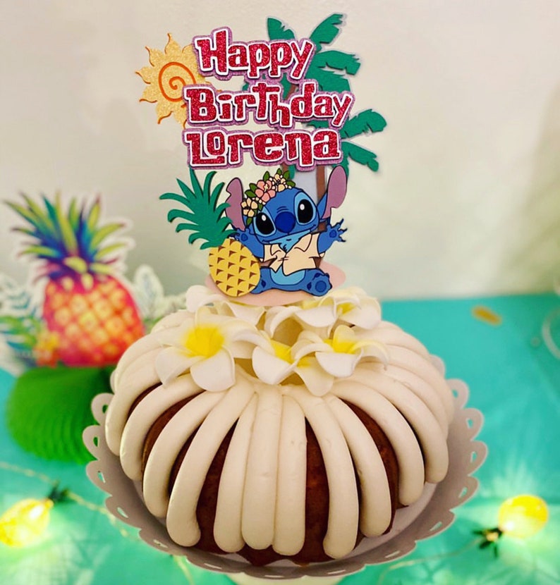 May include: A white cake with white frosting and a blue Stitch character cake topper. The cake topper also includes a pineapple, palm trees, and the text "Happy Birthday Lorena".
