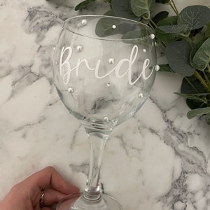 May include: A clear glass with a stem and a round base. The glass has the word "Bride" written on it in white lettering and is decorated with small white pearl-like embellishments.