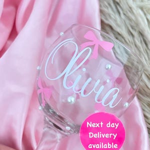 Puede incluir: Un vaso transparente con el nombre "Olivia" escrito en cursiva blanca y lazos rosas. El vaso está decorado con pequeñas cuentas blancas. El vaso está sobre una superficie rosa con una pegatina circular rosa que dice "Entrega al día siguiente disponible".
