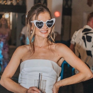 Pu&ograve; includere: Una sposa con un abito bianco senza spalline e occhiali da sole a forma di cuore che recitano "L&A" e "3.00.23".