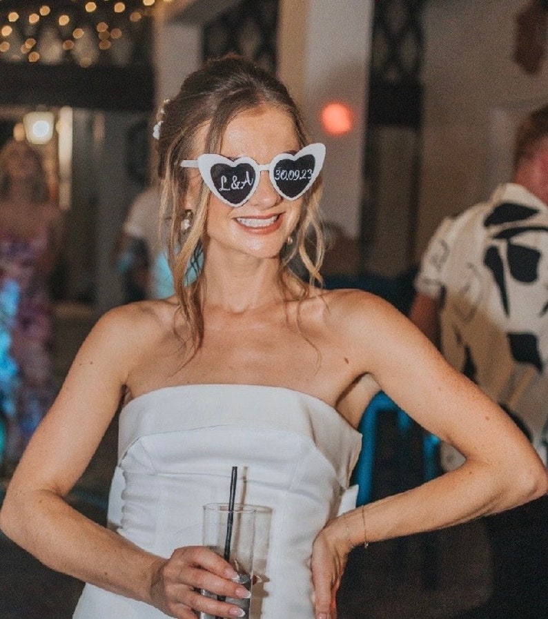 Pu&ograve; includere: Una sposa con un abito bianco senza spalline e occhiali da sole a forma di cuore che recitano "L&A" e "3.00.23".