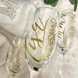 May include: Three clear glass champagne flutes with gold lettering. The flutes are decorated with the words "Bride", "Bridesmaid", and "Sister in Law".