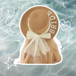 May include: A tan straw sun hat with the word "BRIDE" spelled out in white letters. The hat has a white tulle bow and is decorated with pearls. The hat is designed for a bride.