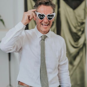 Pu&ograve; includere: Un uomo con una camicia bianca abbottonata e una cravatta grigia sorride e tiene un paio di occhiali da sole bianchi a forma di cuore con il testo "L&A 30.09.23" scritto su di essi.
