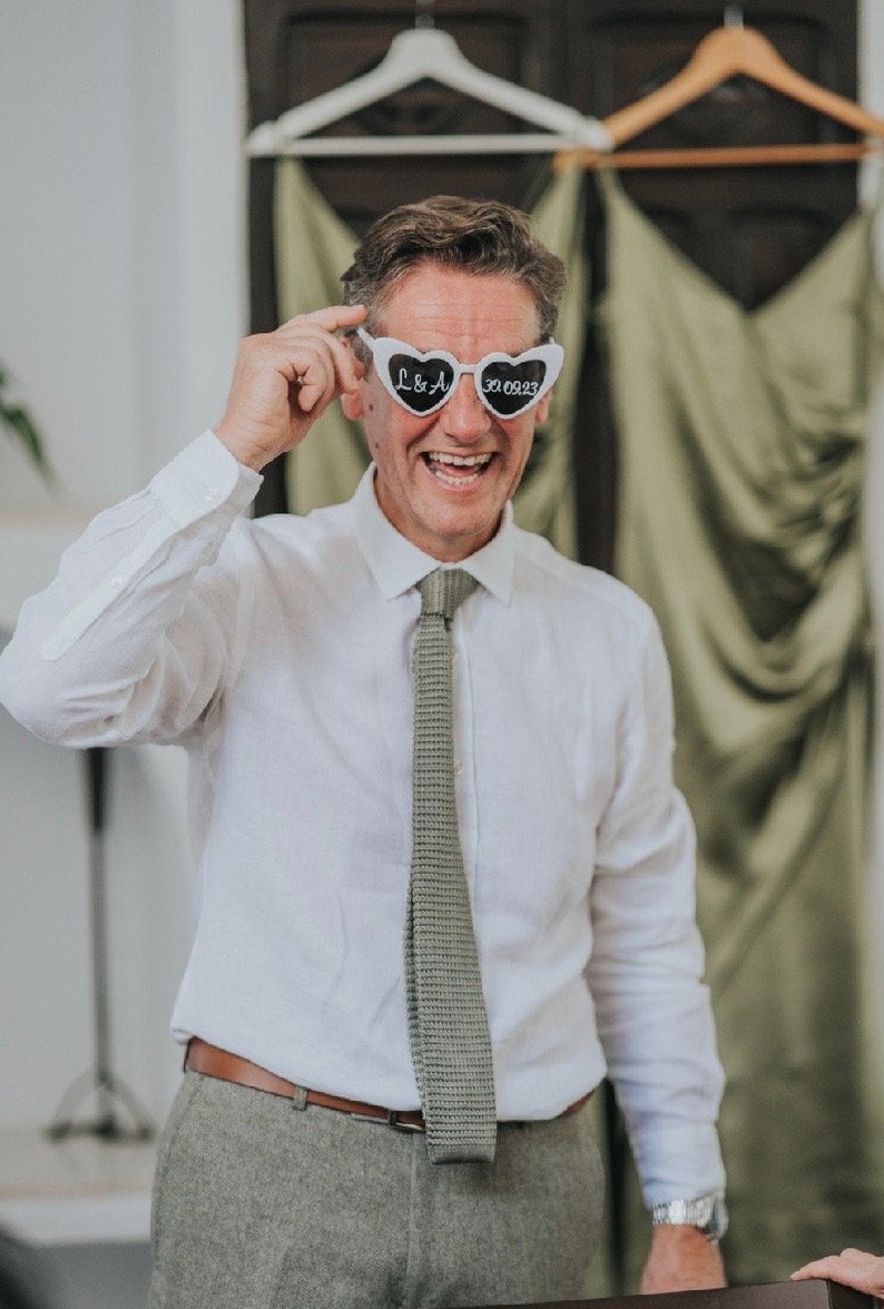 Pu&ograve; includere: Un uomo con una camicia bianca abbottonata e una cravatta grigia sorride e tiene un paio di occhiali da sole bianchi a forma di cuore con il testo "L&A 30.09.23" scritto su di essi.