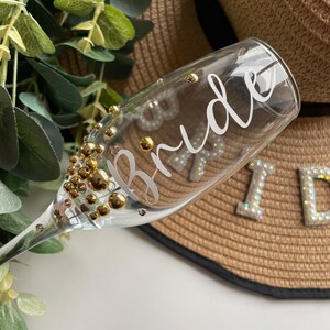 May include: A clear glass champagne flute with gold glitter and the word "Bride" written on it.